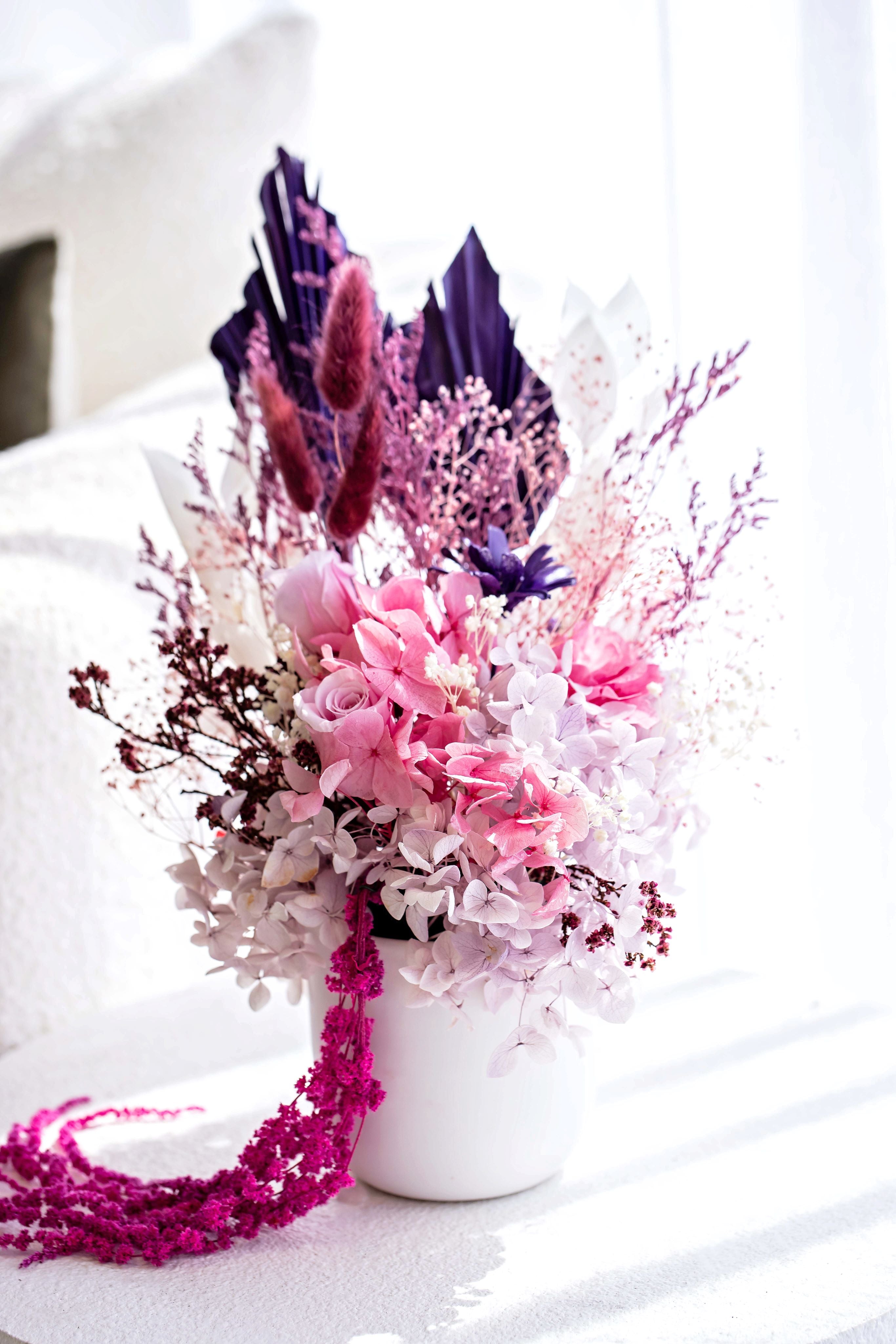 Preserved flower arrangement in a white ceramic pot featuring soft purples, whites, and bright pinks. The subtle blush creates a calming effect, complementing the serenity of any tranquil space. Available in three sizes