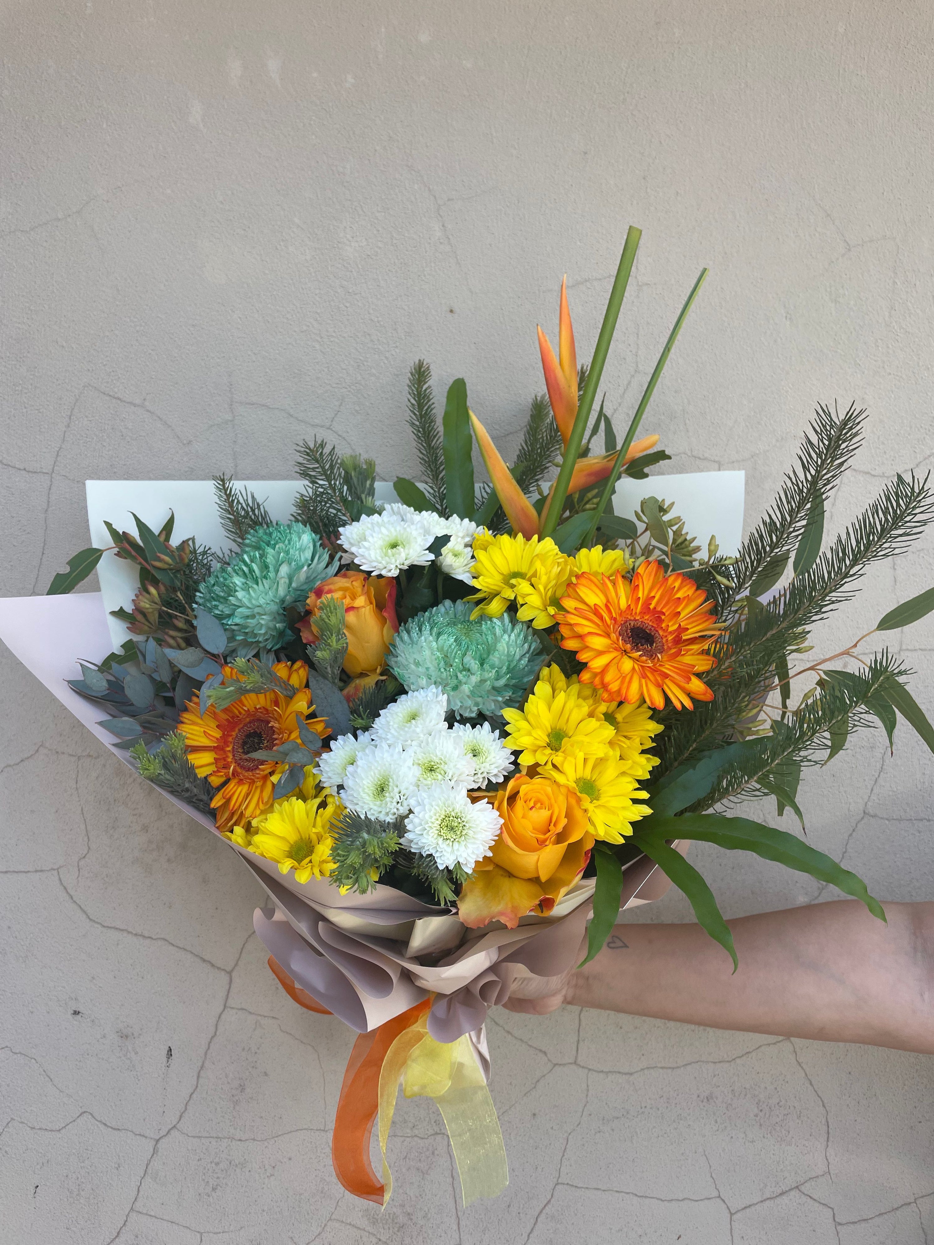 Bouquet of flowers with orange, yellow, and green colors held by a hand on a textured surface. The perfect bouquet for same day delivery accross Prrth from The Flower Boutique Ashby 6065