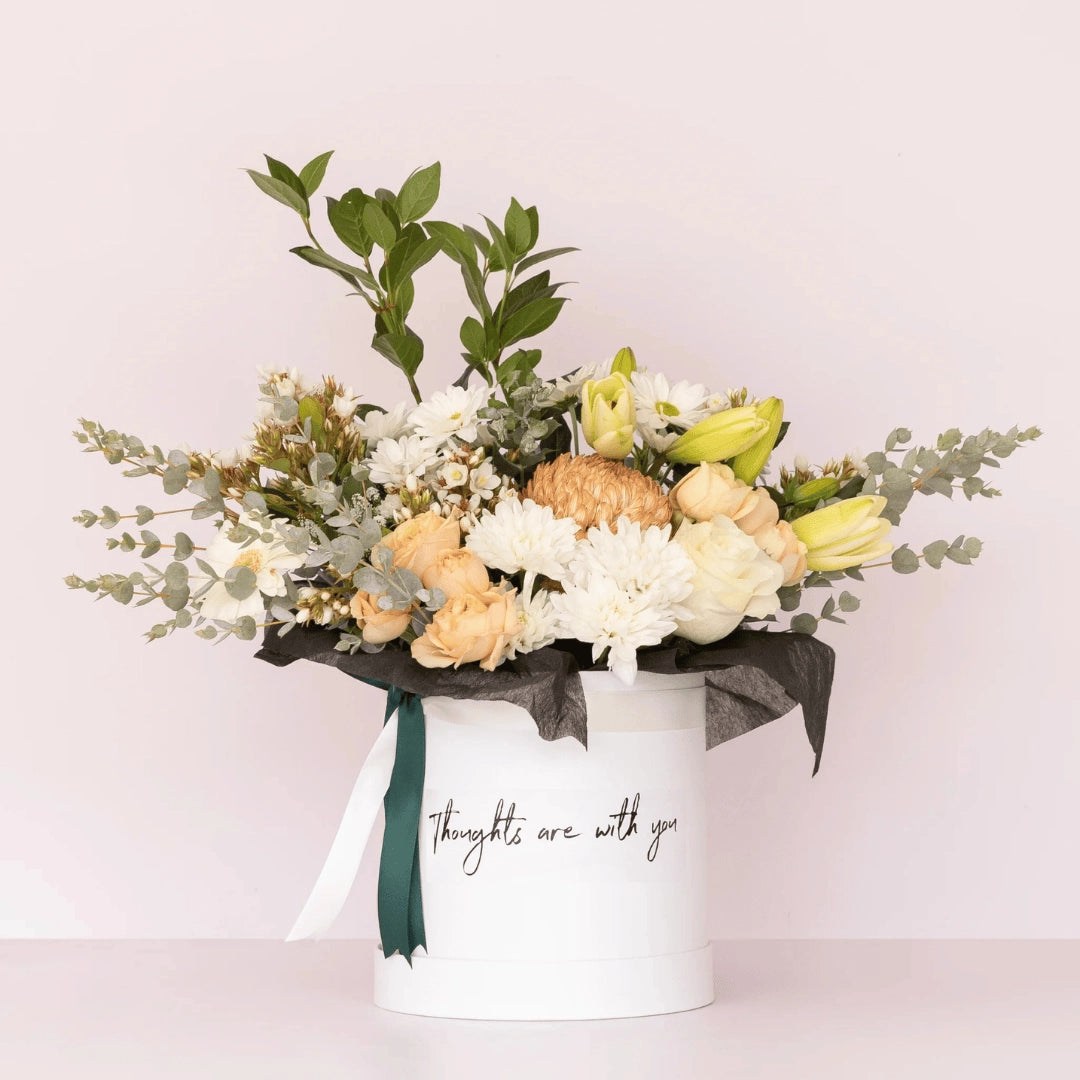 A Deluxe flower box with soft & neutral blooms inside a white hatbox with the message "thoughts are with you" printed on the front of the box available for Flower Delivery Perth | Sympathy Flowers | Funeral Flowers