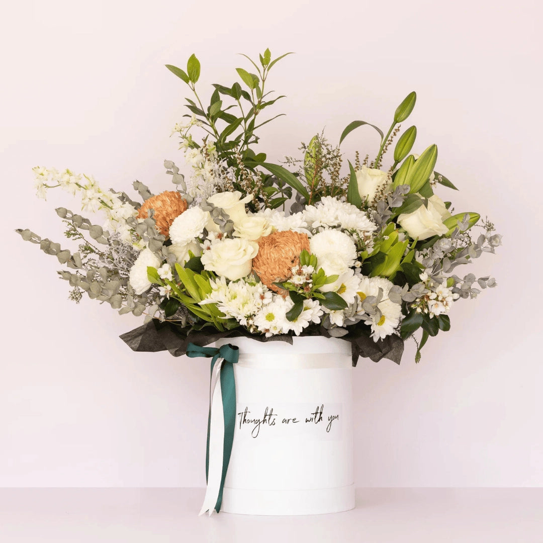 A Lavish flower box with soft & neutral blooms inside a white hatbox with the message "thoughts are with you" printed on the front of the box available for Flower Delivery Perth | Sympathy Flowers | Funeral Flowers