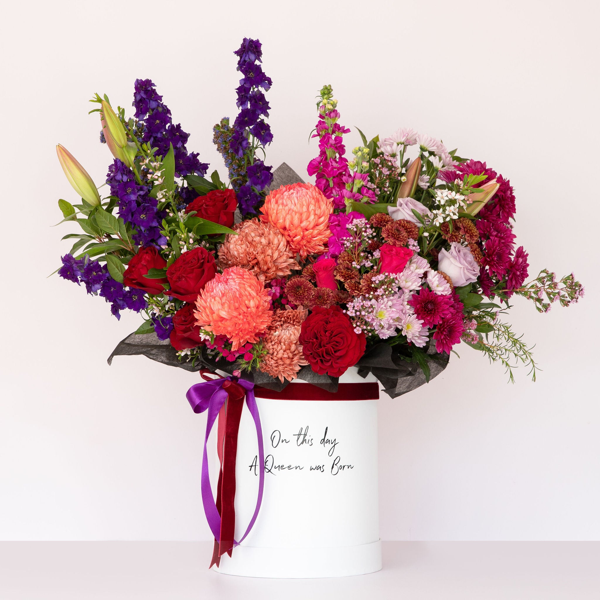 A Lavish flower box with bold & berry flower colours, purples, reds, pinks inside a white keepsake hatbox which is reusable and has ribbon around it with a bow and a print on the front of the box that reads "on this day a Queen was born" a great Happy Birthday gift available for delivery across Perth from The Flower Boutique expert premium florist in Ashby on Pinjar Road