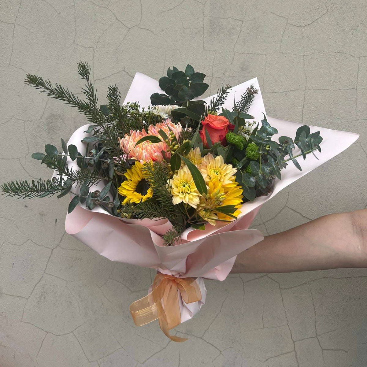 Bright Posy bouquet with yellow, orange and peach flowers and green foliage, held in pale pink wrapping with ribbon