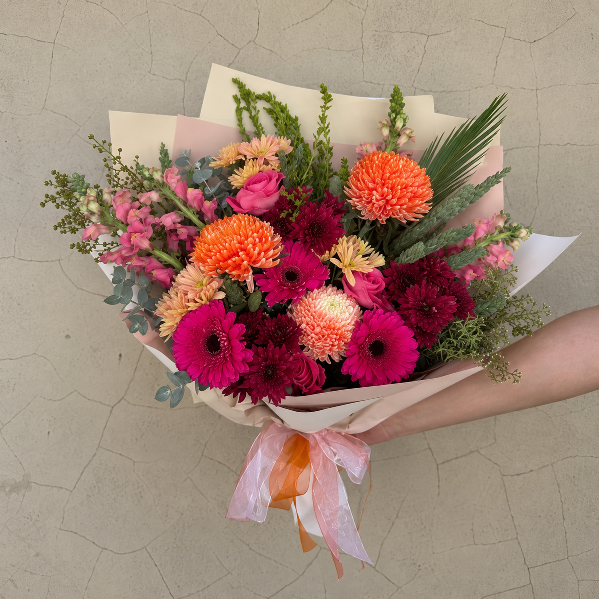 Cherry Spice bouquet with hot pink gerberas, orange chrysanthemums and burgundy flowers, held against a beige wall