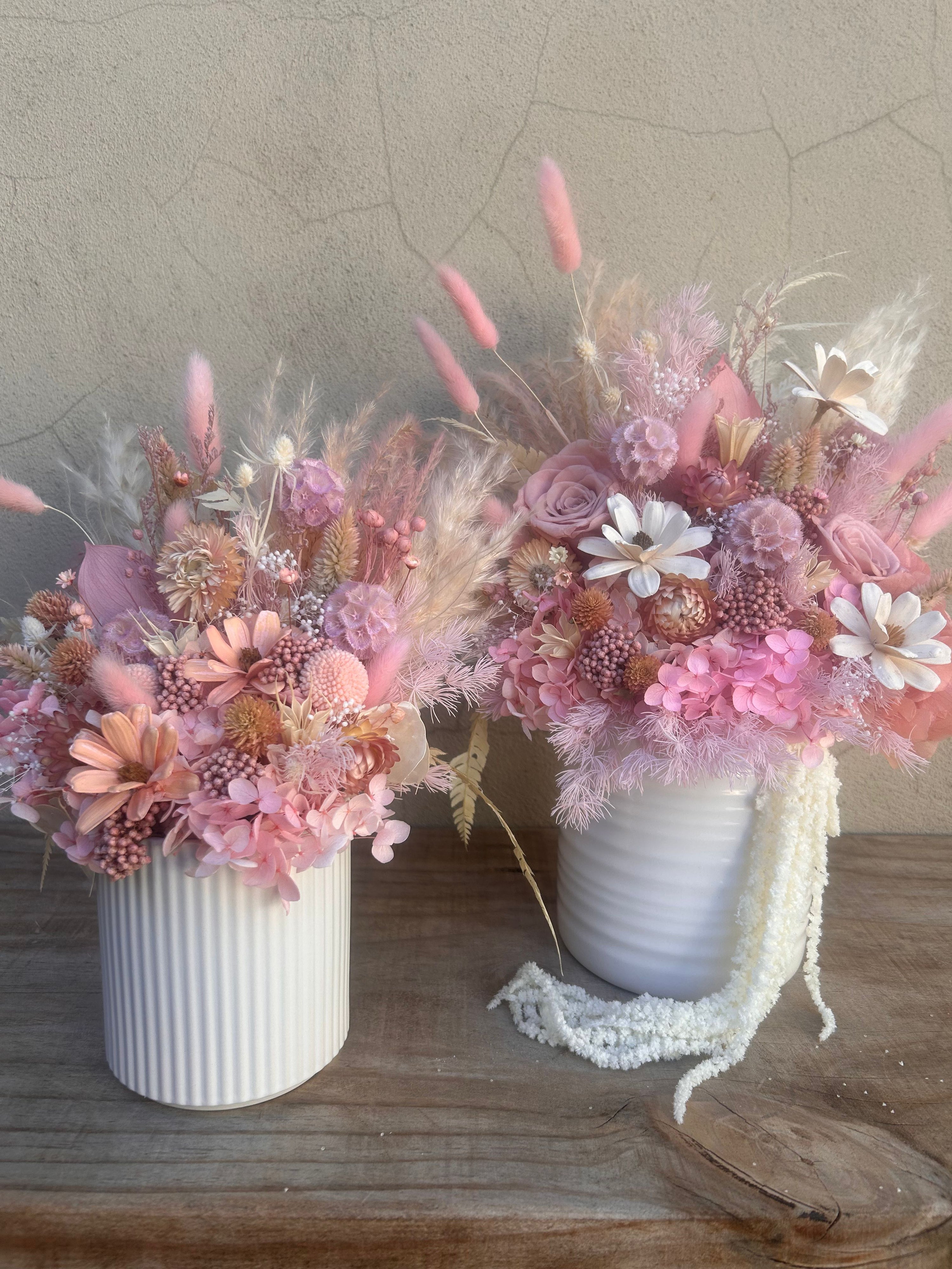 Two white ribbed ceramic vases filled with a dried floral arrangement in shades of pink, peach, cream, and white, displayed on a wooden surface against a textured beige wall.