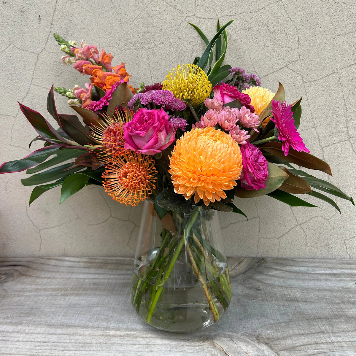 Festa Floreale arrangement with hot pink roses, orange chrysanthemums and textured foliage in a clear glass vase