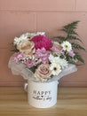 Mother’s Day flower arrangement in a white round box with 'Happy Mother’s Day' printed on it, featuring pastel pink roses, white chrysanthemums, soft pink daisies, and a vibrant hot pink bloom, styled with delicate greenery. Created by The Flower Boutique Perth.