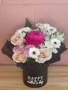 Mother’s Day flower arrangement in a black round box with 'Happy Mother’s Day' printed on it, featuring pastel pink roses, white chrysanthemums, soft pink daisies, and a vibrant hot pink bloom, styled with delicate greenery. Created by The Flower Boutique Perth.