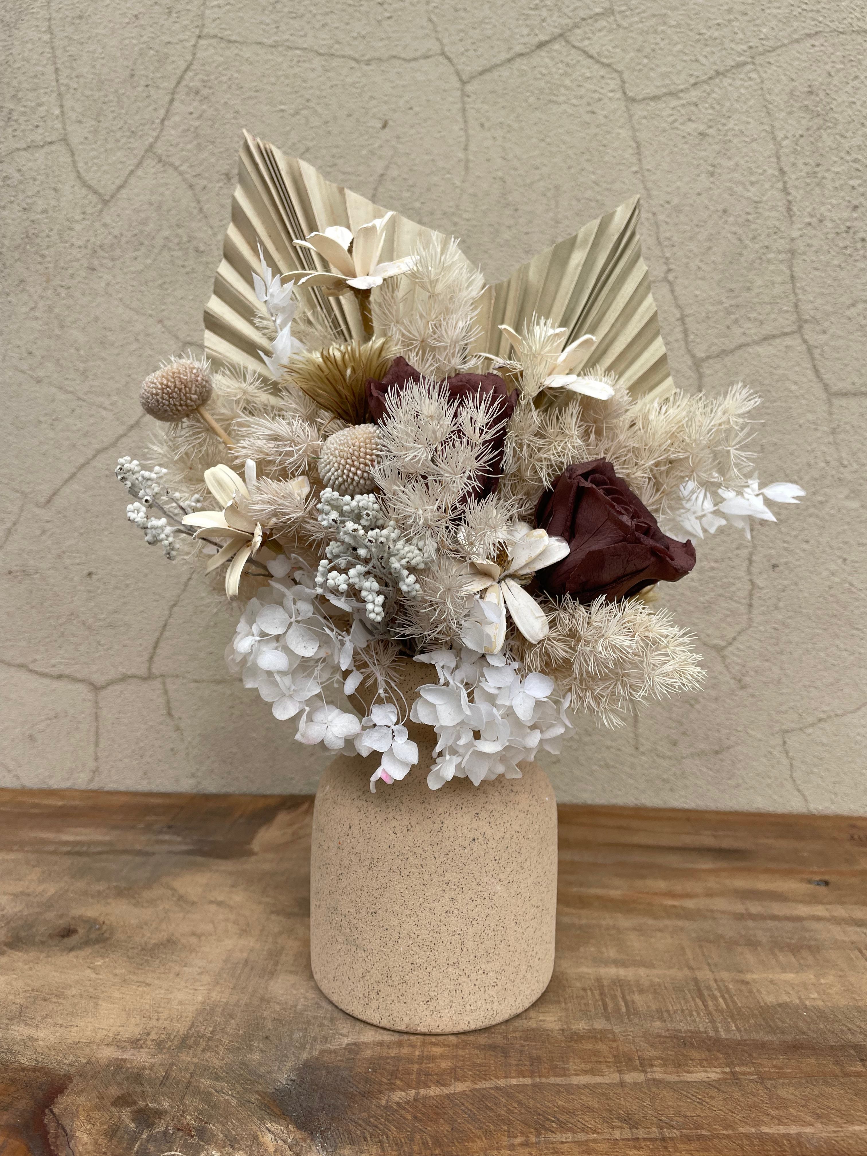 Dried floral arrangement with preserved roses, white hydrangeas, palm spears, and textured fillers in a stone ceramic vase.