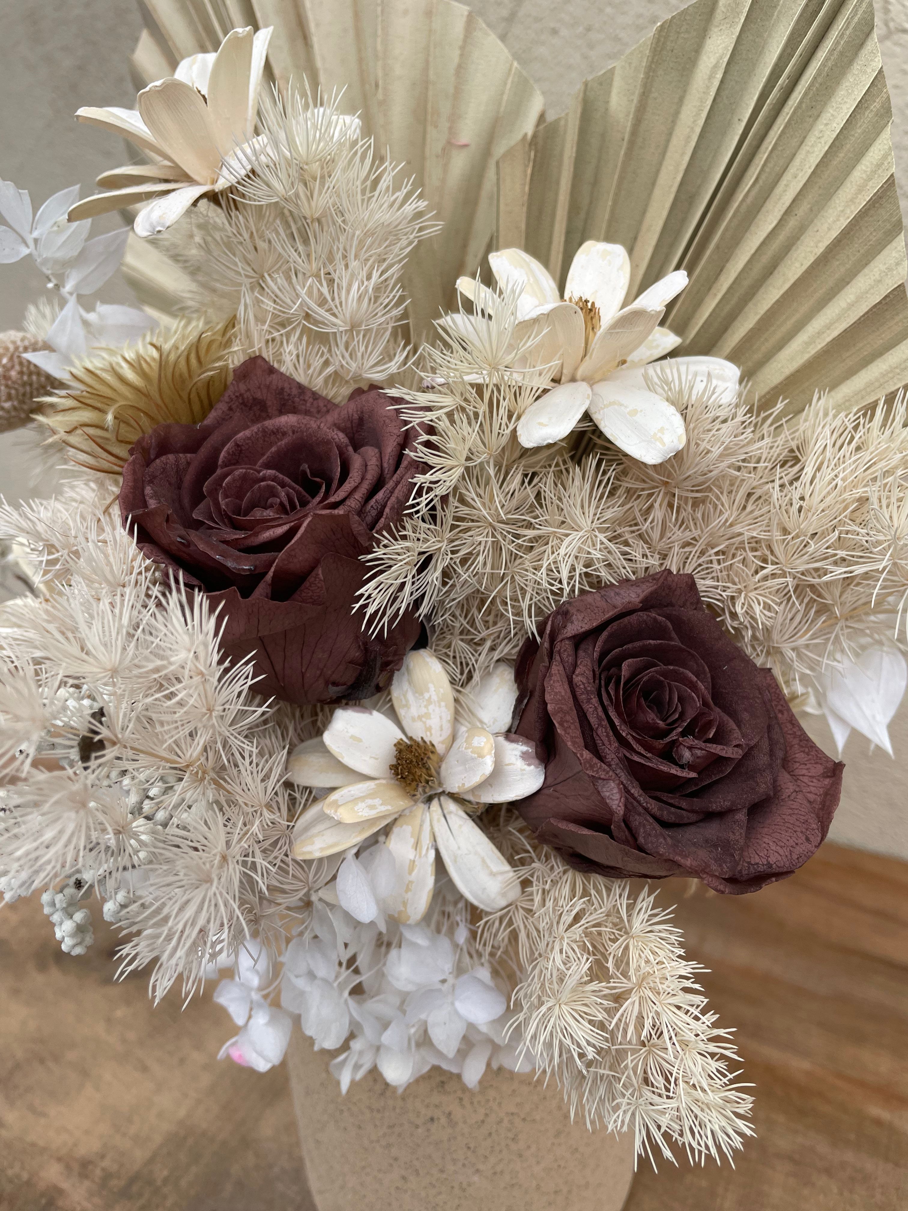 Dried floral arrangement with preserved roses, white hydrangeas, palm spears, and textured fillers in a stone ceramic vase.