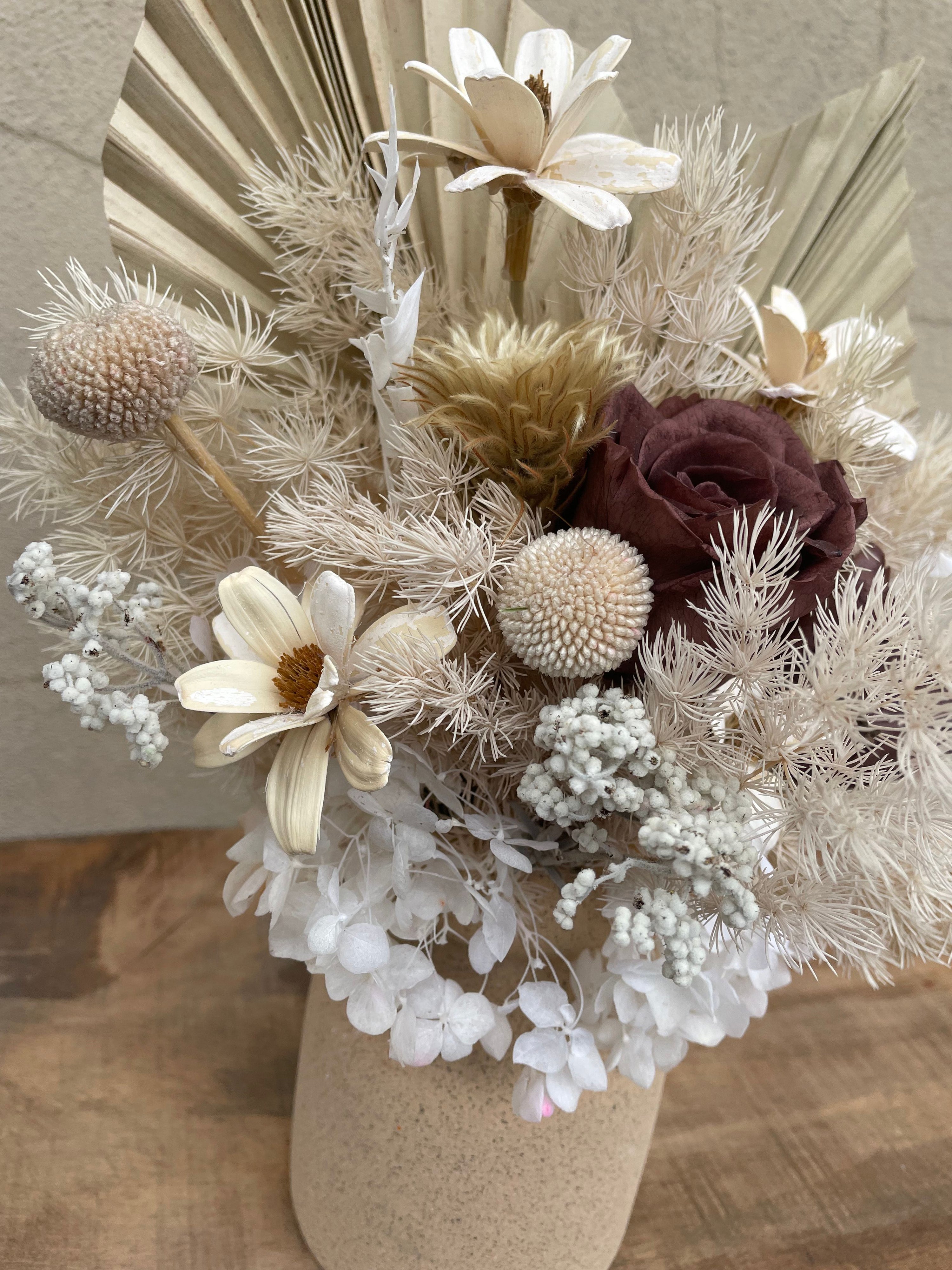 Dried floral arrangement with preserved roses, white hydrangeas, palm spears, and textured fillers in a stone ceramic vase.