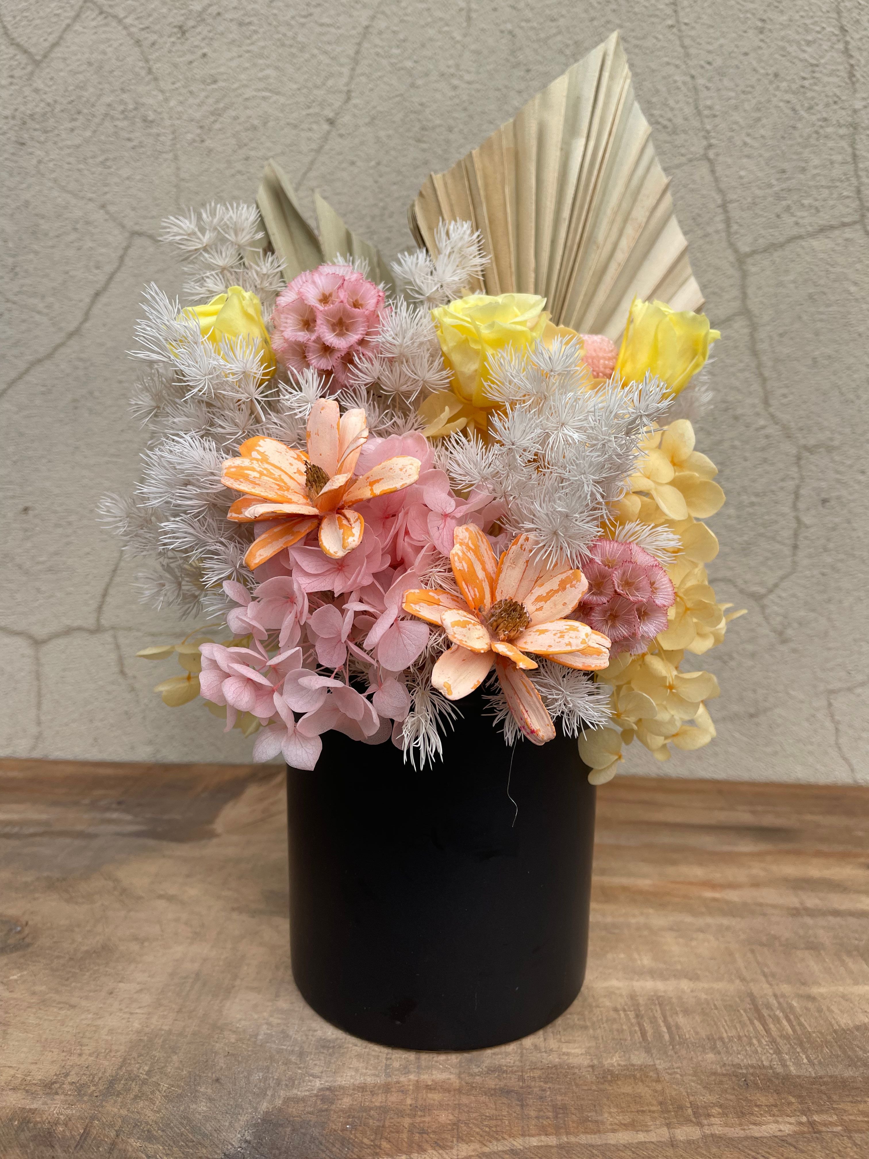 Preserved floral arrangement in a matte black ceramic pot featuring yellow roses, blush pink hydrangeas, peach daisies, and textured dried fillers with palms.
