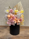 Preserved floral arrangement in a matte black ceramic pot featuring yellow roses, blush pink hydrangeas, peach daisies, and textured dried fillers with palms.