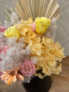 Preserved floral arrangement in a matte black ceramic pot featuring yellow roses, blush pink hydrangeas, peach daisies, and textured dried fillers with palms.