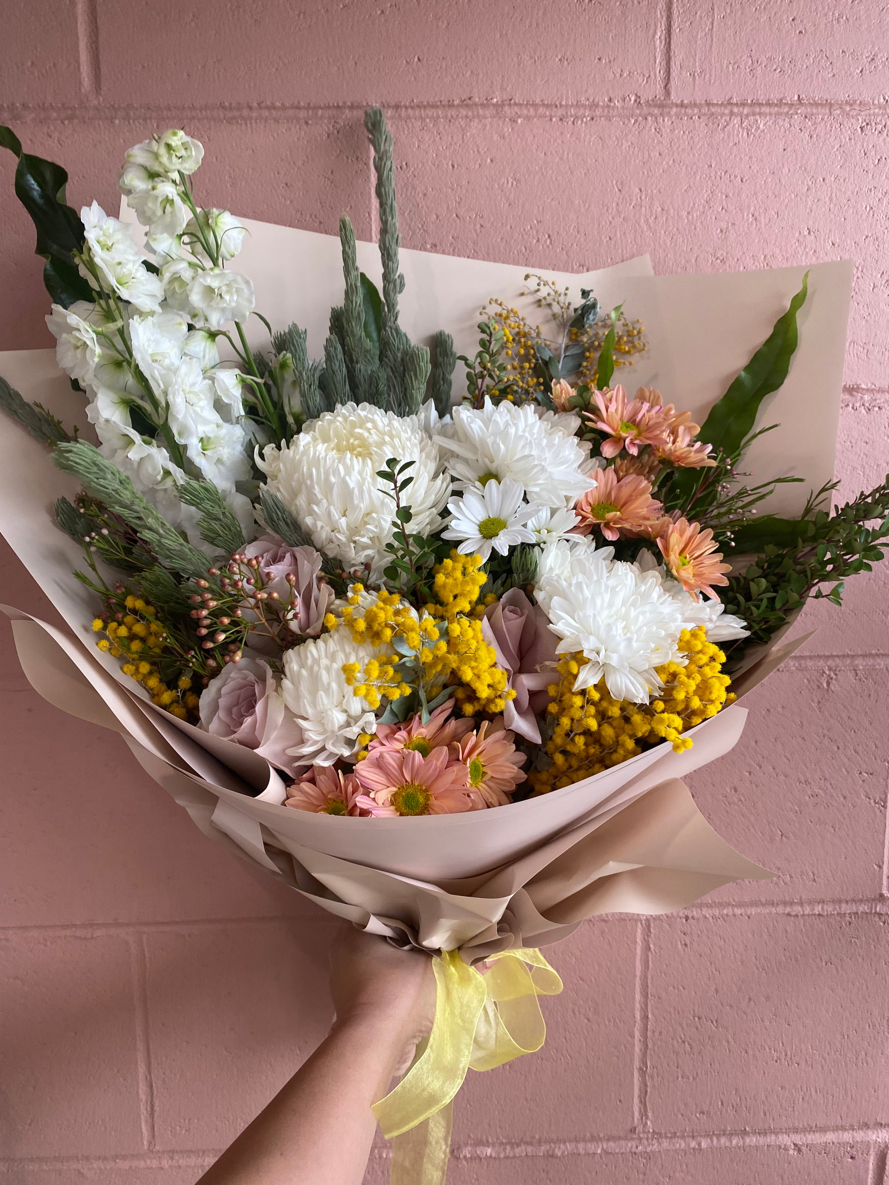 A hand-tied fresh flower bouquet called Sunlit Dawn, featuring yellow wattle, soft pink chrysanthemums, white daisies, pastel roses, and lush greenery, wrapped in soft paper with a yellow ribbon — available for Perth delivery.