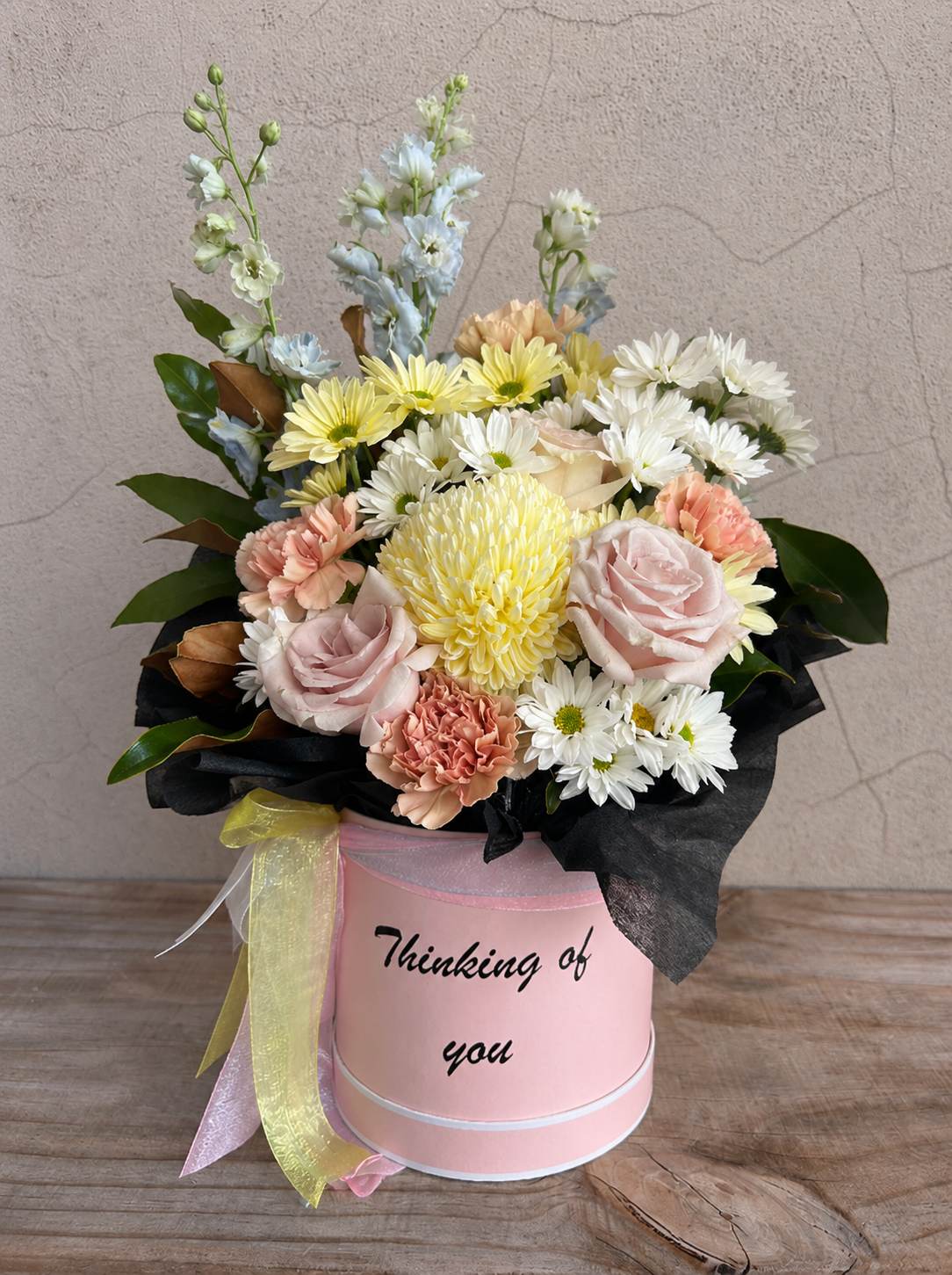 Soft pastel flower arrangement in a pink hatbox with blush roses, white daisies, peach carnations and yellow chrysanthemum, styled on a rustic wooden table against a textured neutral wall.